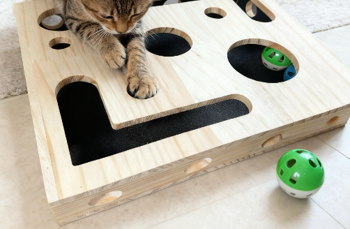 Cat playing with wooden puzzle feeder box, paw reaching into treat hole, with bonus rattle ball